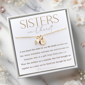 May include: A white box with a gold necklace inside. The necklace has a small pearl charm and a gold charm with the letter "S" on it. The box has a white background with black text that reads "SISTERS in Christ" and a quote about friendship.