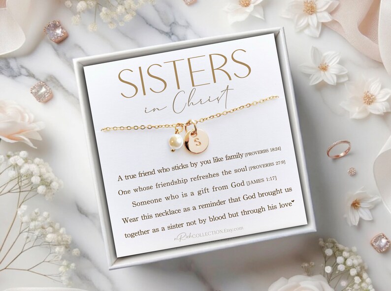 May include: A white box with a gold necklace inside. The necklace has a small pearl charm and a gold charm with the letter "S" on it. The box has a white background with black text that reads "SISTERS in Christ" and a quote about friendship.