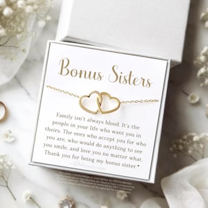 May include: A gold-colored necklace with two connected heart pendants, presented in a white gift box. The box features the words "Bonus Sisters" in gold lettering, along with a heartfelt message. The background includes floral accents.