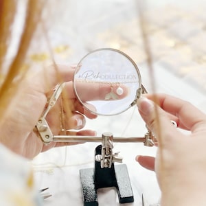 May include: A close-up of a gold chain necklace with a small white pearl and a gold mermaid tail charm. The necklace is being held by a person's hand and is being viewed through a magnifying glass. The text "nRich COLLECTION" is visible in the background.