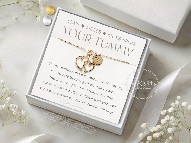 Op de afbeelding: Goudkleurige ketting met twee hartvormige hangers en een kleine ronde bedel, gepresenteerd in een witte geschenkdoos. De kaart binnenin luidt "LOVE KISSES KICKS FROM YOUR TUMMY" met een gedicht. Een sentimenteel cadeau.