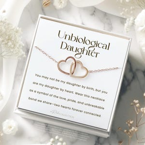 May include: A white card with the text "Unbiological Daughter" and a message about a necklace with two intertwined hearts. The card is sitting on a white surface with white flowers.