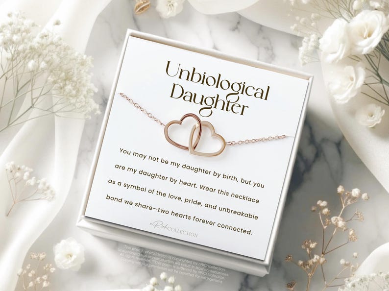 May include: A white card with the text "Unbiological Daughter" and a message about a necklace with two intertwined hearts. The card is sitting on a white surface with white flowers.
