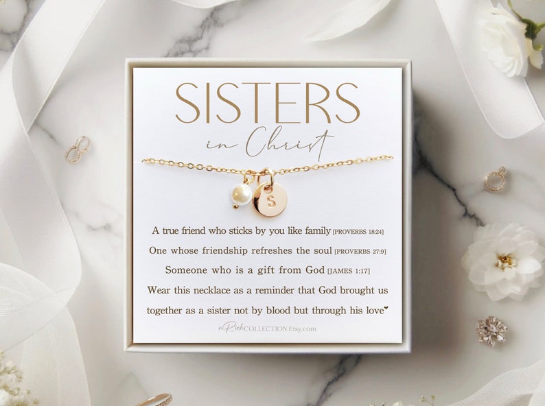 May include: A white box with a gold necklace inside. The necklace has a pearl charm and a gold charm with the letter "S". The box has a white background with the text "SISTERS in Christ" in gold lettering. The text below the necklace reads: "A true friend who sticks by you like family [PROVERBS 18:24] One whose friendship refreshes the soul [PROVERBS 27:9] Someone who is a gift from God [JAMES 1:17] Wear this necklace as a reminder that God brought us together as a sister not by blood but through his love."