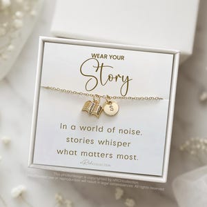 Może przedstawiać: Złota bransoletka z zawieszką w kształcie książki i inicjału, prezentowana w białym pudełku. Karta w środku głosi "WEAR YOUR Story" i cytat o historiach. Tło obejmuje białe kwiaty i pierścionek.