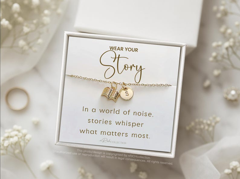 Może przedstawiać: Złota bransoletka z zawieszką w kształcie książki i inicjału, prezentowana w białym pudełku. Karta w środku głosi "WEAR YOUR Story" i cytat o historiach. Tło obejmuje białe kwiaty i pierścionek.