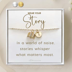 Może przedstawiać: Złota bransoletka z zawieszką w kształcie książki i inicjału, prezentowana na białej karcie. Na karcie napis "WEAR YOUR Story" i cytat o historiach. Tło zawiera wstążkę i kwiaty.