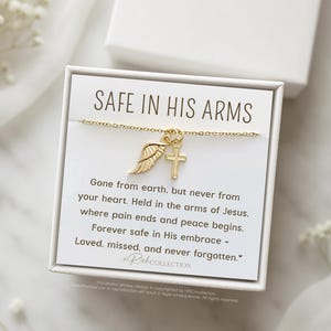 May include: A white card with gold lettering that reads "Safe in His Arms". The card features a gold chain necklace with a small gold wing and cross charm. The card also includes a message about being safe in the arms of Jesus.