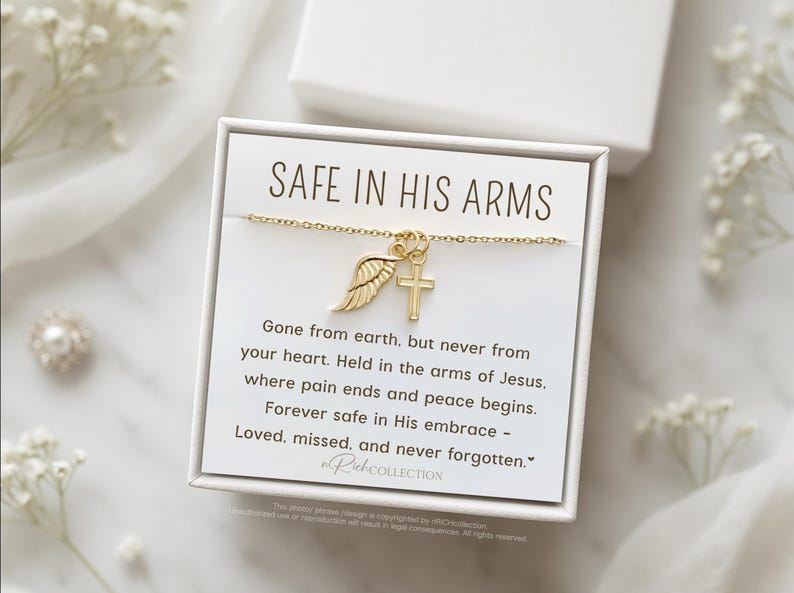 May include: A white card with gold lettering that reads "Safe in His Arms". The card features a gold chain necklace with a small gold wing and cross charm. The card also includes a message about being safe in the arms of Jesus.