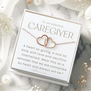 May include: A white card with gold lettering reads "To an amazing caregiver." The card features a gold necklace with two intertwined hearts. The text on the card reads: "A heart so giving, a soul so kind, with love and sacrifice intertwined. Wear this as a reminder that we are connected by heart and always will be."