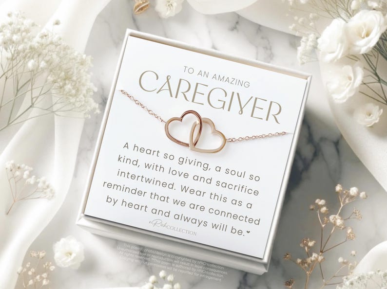 May include: A white card with gold lettering reads "To an amazing caregiver." The card features a gold necklace with two intertwined hearts. The text on the card reads: "A heart so giving, a soul so kind, with love and sacrifice intertwined. Wear this as a reminder that we are connected by heart and always will be."
