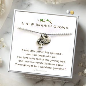 May include: A silver necklace with a tree pendant and a small round charm, presented in a white box. The box has text that reads "A New Branch Grows" and a message about family. The background includes white flowers and a marble-like surface.