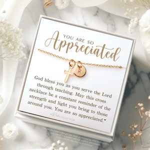 May include: A white gift box with a gold necklace inside. The necklace has a small cross charm and a round charm with the letter "R" on it. The box has a message that reads "You are so appreciated. God bless you as you serve the Lord through teaching. May this cross necklace be a constant reminder of the strength and light you bring to those around you. You are so appreciated."