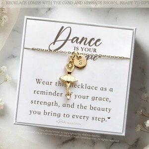 May include: A white gift box with a gold necklace featuring a ballerina charm and a letter 'S' charm. The box includes a card with the text "Dance is your spirit. Wear this necklace as a reminder of your grace, strength, and the beauty you bring to every step."