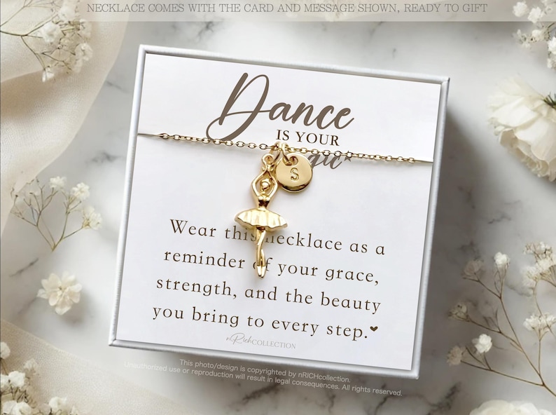 May include: A white gift box with a gold necklace featuring a ballerina charm and a letter 'S' charm. The box includes a card with the text "Dance is your spirit. Wear this necklace as a reminder of your grace, strength, and the beauty you bring to every step."