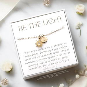 May include: A gold necklace with a sun and a circle charm. The necklace comes in a white box with the words "Be the Light" printed on it. The box also includes a message about wearing the necklace as a reminder to shine bright.