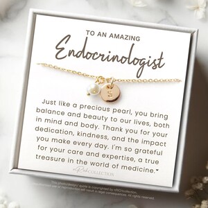 May include: A gold-colored necklace with a small pearl and a round charm with the letter "S" is displayed on a white card inside a white box. The card reads "TO AN AMAZING Endocrinologist".