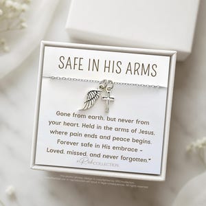 May include: A white card with the text "SAFE IN HIS ARMS" and a silver necklace with a cross and angel wing charm. The card reads "Gone from earth, but never from your heart. Held in the arms of Jesus. Where pain ends and peace begins. Forever safe in His embrace - Loved, missed, and never forgotten."