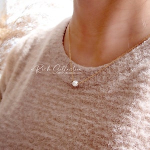 K&ouml;nnte beinhalten: Eine zarte goldene Kettenkette mit einem einzelnen wei&szlig;en Perlenanh&auml;nger. Die Kette wird von einer Person getragen, die einen hellbraunen Pullover tr&auml;gt.