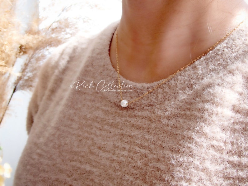 K&ouml;nnte beinhalten: Eine zarte goldene Kettenkette mit einem einzelnen wei&szlig;en Perlenanh&auml;nger. Die Kette wird von einer Person getragen, die einen hellbraunen Pullover tr&auml;gt.
