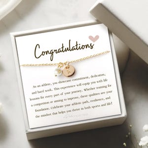 May include: A white gift box with a gold necklace inside. The necklace has a small gold charm with a letter on it. The box has a pink heart and the text "Congratulations" on the front. The text inside the box reads "As an athlete, you showcase commitment, dedication, and hard work. This experience will equip you with life lessons for every part of your journey. Whether training for a competition or aiming to improve, these qualities are your foundation. Celebrate your athletic path, resilience, and the mindset that helps you thrive in both sports and life!"