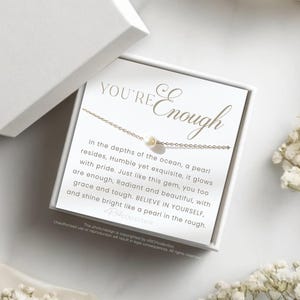 May include: A white box containing a gold necklace with a pearl charm. The box has the text "YOU'RE Enough" on the front. The text inside the box reads "In the depths of the ocean, a pearl resides, Humble yet exquisite, it glows with pride. Just like this gem, you too are enough, Radiant and beautiful, with grace and tough. BELIEVE IN YOURSELF, and shine bright like a pearl in the rough."