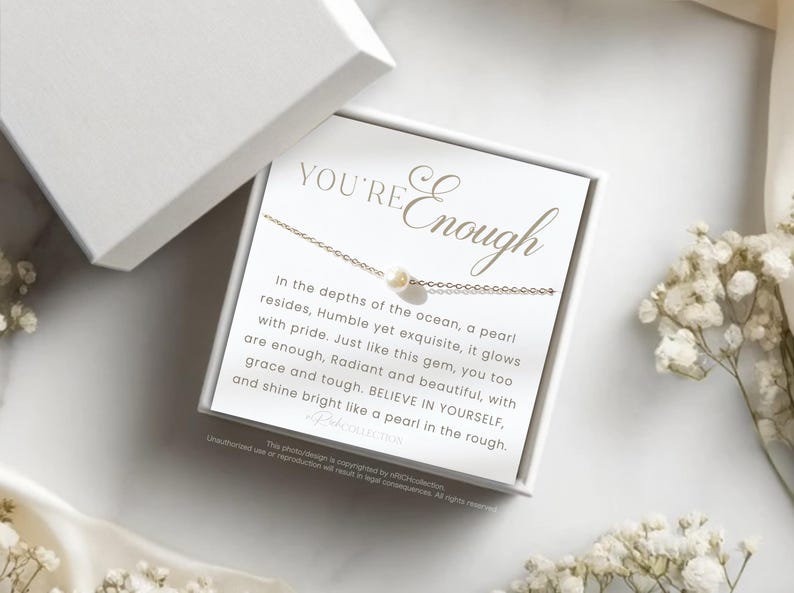 May include: A white box containing a gold necklace with a pearl charm. The box has the text "YOU'RE Enough" on the front. The text inside the box reads "In the depths of the ocean, a pearl resides, Humble yet exquisite, it glows with pride. Just like this gem, you too are enough, Radiant and beautiful, with grace and tough. BELIEVE IN YOURSELF, and shine bright like a pearl in the rough."
