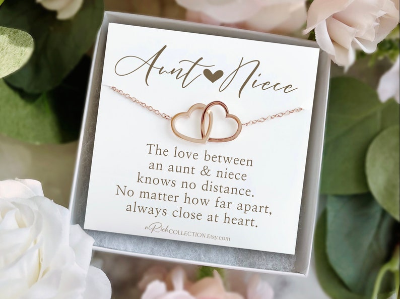 May include: A rose gold necklace with two interlocking heart pendants, presented on a white card inside a gift box. The card reads "Aunt & Niece" and a sentimental message about the bond between them. The background features soft floral accents.