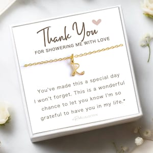 May include: A gold-colored necklace with the letter "R" pendant, presented in a white gift box. The box features the text "Thank You" and a message expressing gratitude. White flowers and greenery surround the box.