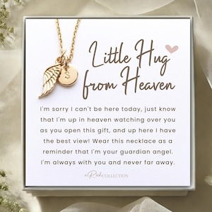 May include: A white card with gold lettering that reads "Little Hug from Heaven". The card is next to a gold necklace with a small wing charm and a round charm. The card includes a message about a guardian angel and a reminder that someone is always with you.