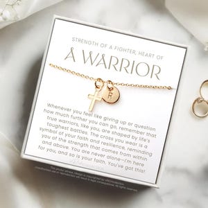 May include: A gold necklace with a cross charm and a letter charm. The necklace is in a white box with the words "Strength of a fighter, heart of a warrior" printed on it. The box also includes an inspirational message about faith and resilience.