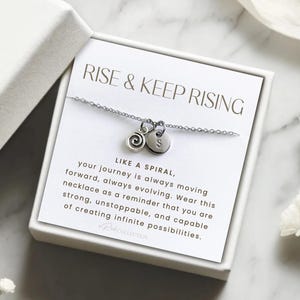 May include: A silver necklace with a spiral charm and a letter charm in a white box. The box has the text "RISE & KEEP RISING" and a message about the necklace being a reminder to be strong and capable of creating infinite possibilities.