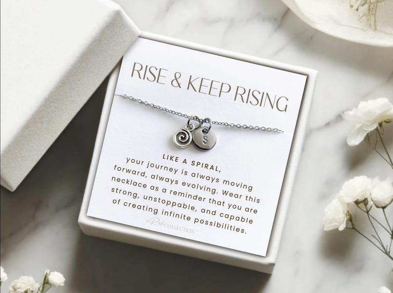 May include: A silver necklace with a spiral charm and a letter charm in a white box. The box has the text "RISE & KEEP RISING" and a message about the necklace being a reminder to be strong and capable of creating infinite possibilities.