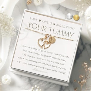 Op de afbeelding: Een goudkleurige ketting met twee hartvormige hangers en een kleine ronde bedel, gepresenteerd op een witte kaart met de tekst "LOVE KISSES KICKS FROM YOUR TUMMY". De kaart zit in een witte doos, omringd door witte bloemen.