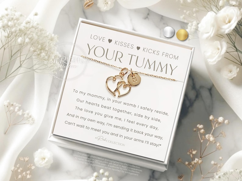 Op de afbeelding: Een goudkleurige ketting met twee hartvormige hangers en een kleine ronde bedel, gepresenteerd op een witte kaart met de tekst "LOVE KISSES KICKS FROM YOUR TUMMY". De kaart zit in een witte doos, omringd door witte bloemen.