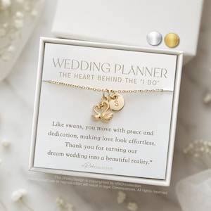 May include: A gold-colored necklace with swan charms and a circular pendant with the letter "S" is displayed on a white card. The card reads "Wedding Planner" and includes text about making love effortless. The necklace is presented in a white box.