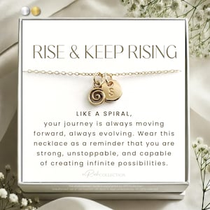 May include: A gold necklace with a spiral charm and a small disc charm with the letter "S" on it. The necklace is in a white box with the text "RISE & KEEP RISING" and a message about the journey being like a spiral.