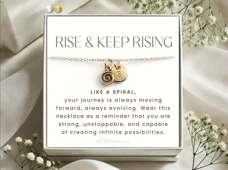 May include: A gold necklace with a spiral charm and a small disc charm with the letter "S" on it. The necklace is in a white box with the text "RISE & KEEP RISING" and a message about the journey being like a spiral.