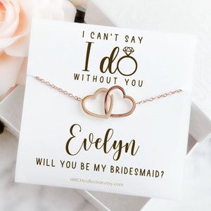 Peut inclure: Une carte blanche avec le texte "Je ne peux pas dire oui sans toi" et une bague en or avec un diamant. Sous le texte se trouve une cha&icirc;ne en or avec deux c&oelig;urs entrelac&eacute;s. La carte dit &eacute;galement "Evelyn, veux-tu &ecirc;tre ma demoiselle d'honneur ?" et inclut une adresse de site Web.
