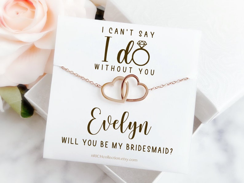 Peut inclure: Une carte blanche avec le texte "Je ne peux pas dire oui sans toi" et une bague en or avec un diamant. Sous le texte se trouve une cha&icirc;ne en or avec deux c&oelig;urs entrelac&eacute;s. La carte dit &eacute;galement "Evelyn, veux-tu &ecirc;tre ma demoiselle d'honneur ?" et inclut une adresse de site Web.