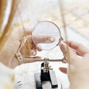 May include: A close-up of a gold chain necklace with a pearl and a mermaid tail charm. The necklace is being held by a pair of hands and is being viewed through a magnifying glass. The magnifying glass has the text "n Rick COLLECTION" printed on it.