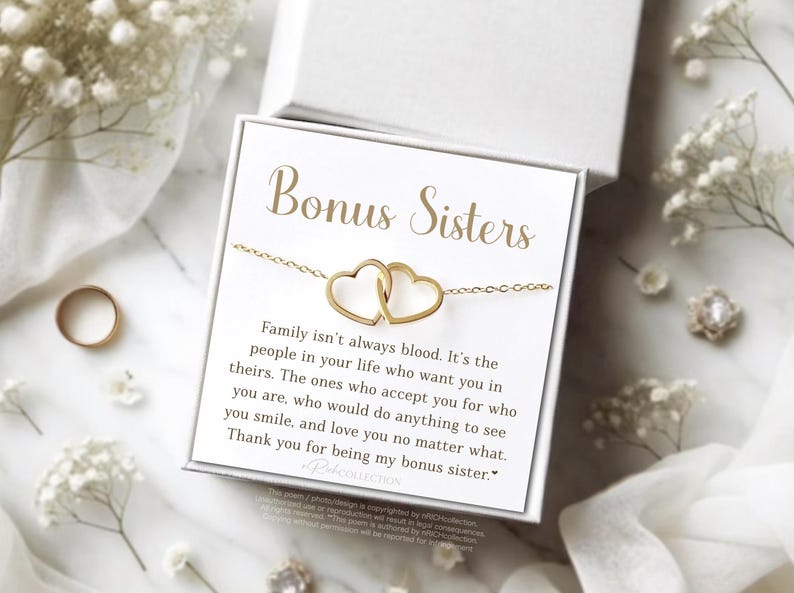 May include: A gold-colored necklace with two connected heart pendants, presented in a white gift box. The box features the words "Bonus Sisters" and a heartfelt message. The background includes white flowers and a gold ring.