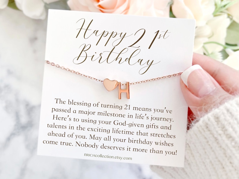 May include: A white card with the text "Happy 21st Birthday" and a message about turning 21. A rose gold necklace with a heart and the letter "H" charm is displayed on the card.