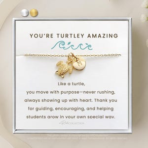 May include: A white card with the text "You're Turtley Amazing" and a gold turtle charm necklace with a wave design. The card also includes a message about moving with purpose and helping students grow.