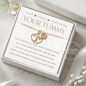 Op de afbeelding: Goudkleurige ketting met twee hartvormige hangers en een kleine ronde bedel, gepresenteerd in een witte geschenkdoos. De kaart binnenin luidt "LOVE KISSES KICKS FROM YOUR TUMMY" met een gedicht. Een sentimenteel cadeau.