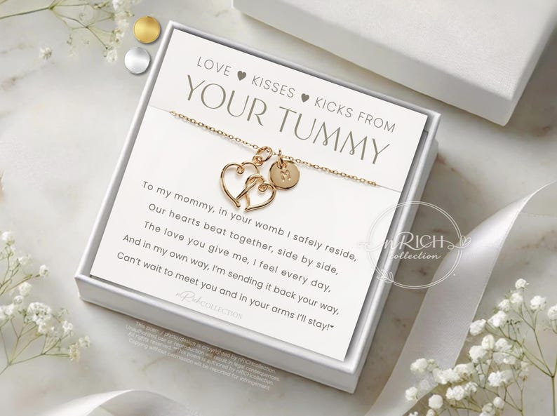 Op de afbeelding: Goudkleurige ketting met twee hartvormige hangers en een kleine ronde bedel, gepresenteerd in een witte geschenkdoos. De kaart binnenin luidt "LOVE KISSES KICKS FROM YOUR TUMMY" met een gedicht. Een sentimenteel cadeau.