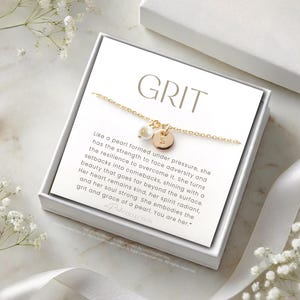 Op de afbeelding: Een witte doos met een gouden kettingketting en een parelbedel. De doos heeft de tekst "GRIT" en een citaat over de kracht en veerkracht van een parel.