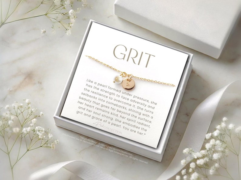 Op de afbeelding: Een witte doos met een gouden kettingketting en een parelbedel. De doos heeft de tekst "GRIT" en een citaat over de kracht en veerkracht van een parel.