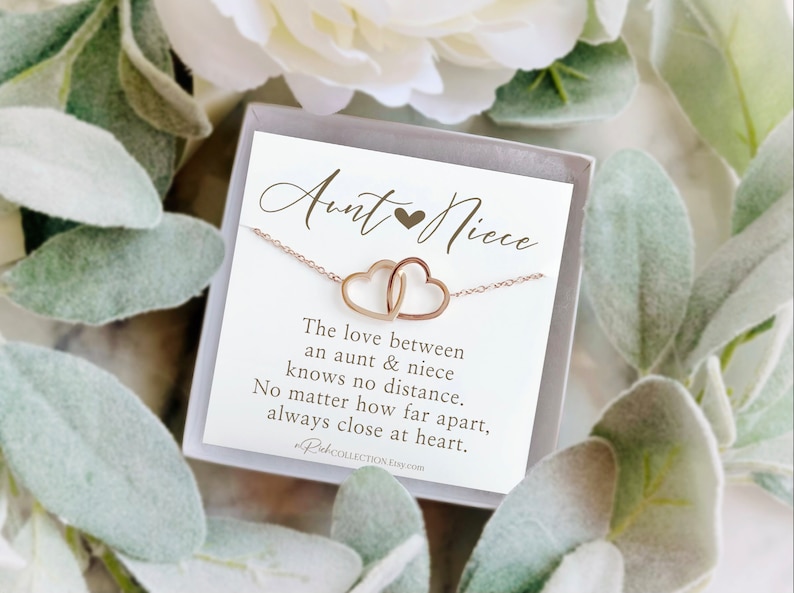 May include: A rose gold necklace featuring two interlocking heart pendants, presented in a gift box with a card. The card reads "Aunt & Niece" and includes a sentimental message about the bond between an aunt and niece.