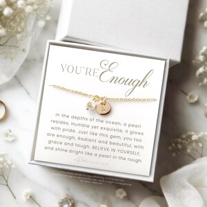 May include: A gold necklace with a pearl and a small gold disc charm. The necklace is displayed on a white card with the text "YOU'RE Enough" and a quote about pearls.
